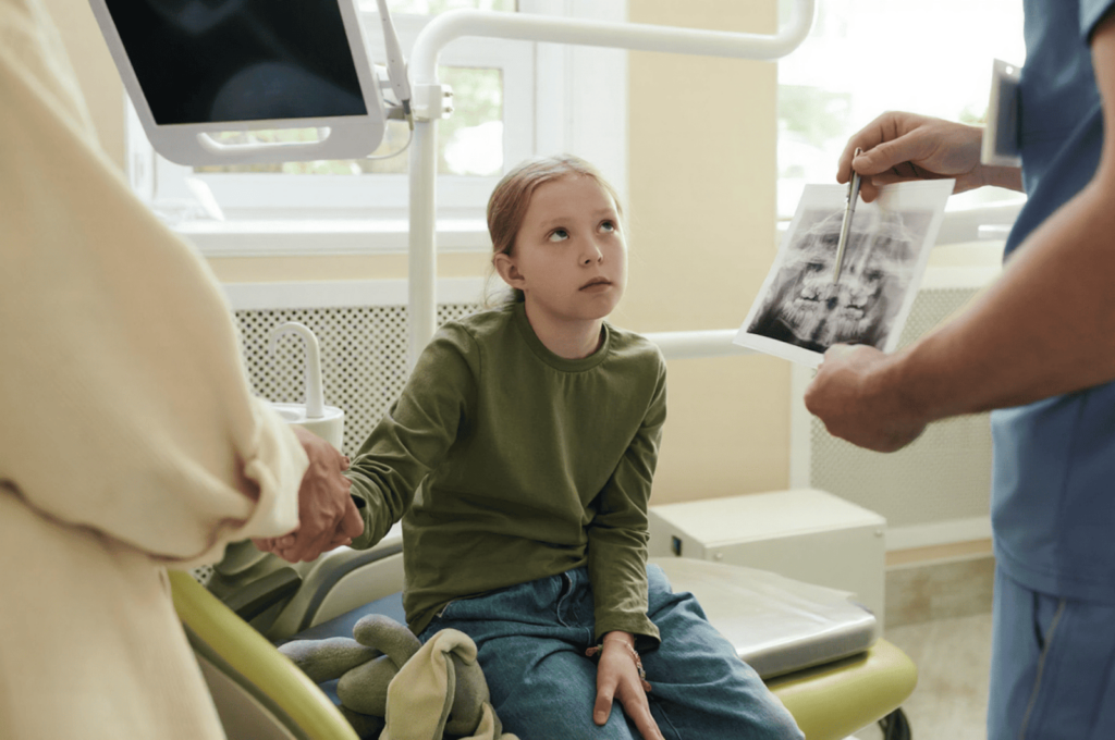 A dentist is pointing at a young girl's oral X-ray and explaining to her mother why he recommends dental crowns over fillings.